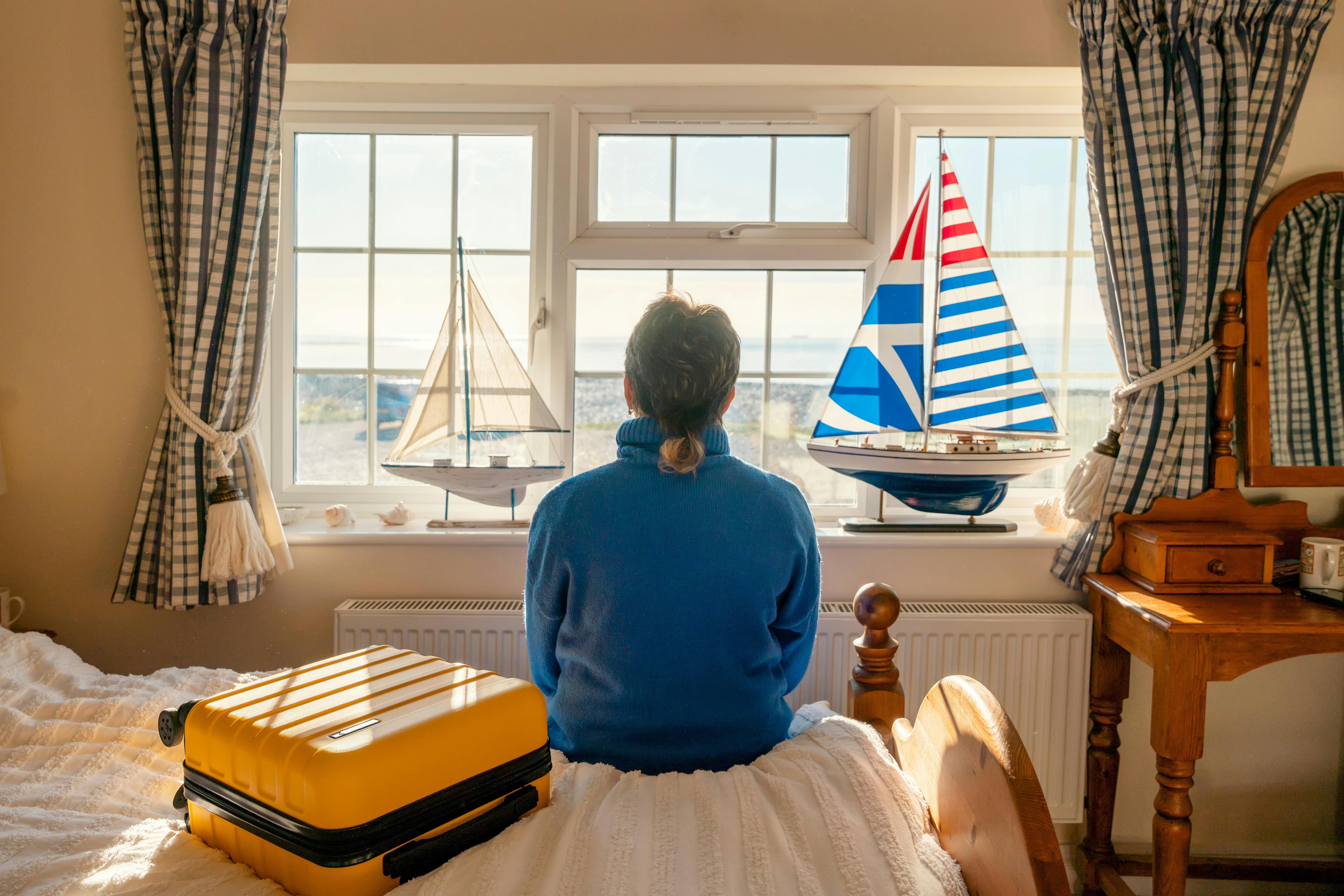 A woman sitting by the window looking out, with a suitcase on her bed, and model ships on the windowsill